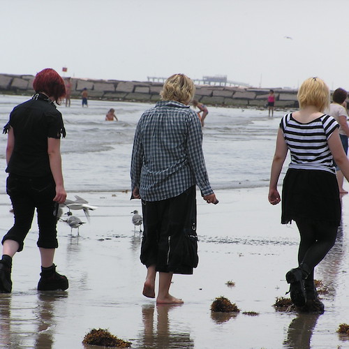 Three friends walk on the beach. One is wearing stylishly ripped all black clothing, another wears a long sleeve plaid shirt with wide-leg capris, and the third is wearing a black and white striped top and black skirt.